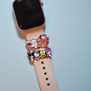 Könnte beinhalten: Eine roségoldene Apple Watch mit einem rosa Armband, verziert mit zwei Sets farbenfroher Charms. Die Charms zeigen Zeichentrickfiguren und Kaffee-Motive. Die Uhr befindet sich vor einem hellblauen Hintergrund.