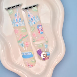 May include: Two clear silicone Apple Watch bands with a colorful print featuring Disney characters, including Mickey Mouse, Minnie Mouse, and Cinderella's castle. The bands are decorated with pastel colors and have a "HUNNY" label on one band.