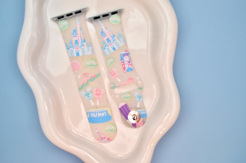 May include: Two clear silicone Apple Watch bands with a colorful print featuring Disney characters, including Mickey Mouse, Minnie Mouse, and Cinderella's castle. The bands are decorated with pastel colors and have a "HUNNY" label on one band.