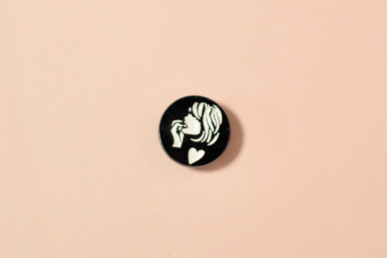 K&ouml;nnte beinhalten: Schwarzer und wei&szlig;er Emaille-Pin mit der Silhouette einer Frau mit kurzen Haaren, die sich auf die Lippe bei&szlig;t und einen herzf&ouml;rmigen Anh&auml;nger tr&auml;gt.