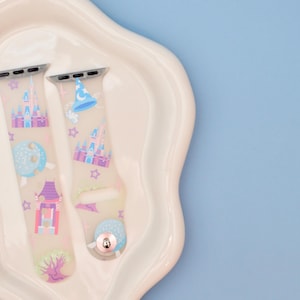 May include: Two pastel-colored silicone Apple Watch bands with a Disney theme. The bands feature images of Cinderella's castle, a magic wand, a Mickey Mouse hat, and other Disney icons.