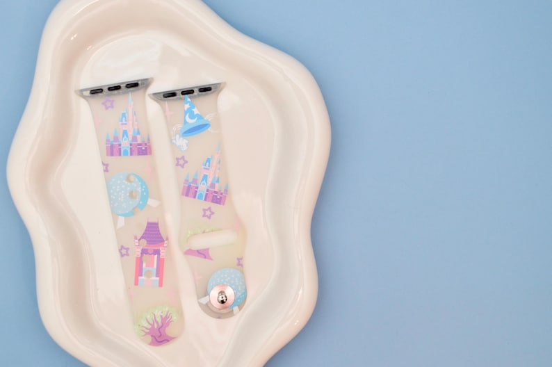 May include: Two pastel-colored silicone Apple Watch bands with a Disney theme. The bands feature images of Cinderella's castle, a magic wand, a Mickey Mouse hat, and other Disney icons.
