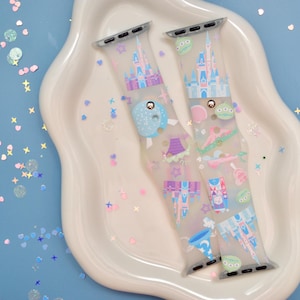 May include: Two clear Apple Watch bands with a colorful Disney theme. The bands feature a pink castle, a blue and white spaceship, and other Disney characters. The bands are on a white plate with a blue background.