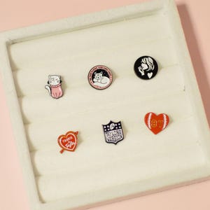 K&ouml;nnte beinhalten: Ein wei&szlig;es Tablett mit sechs Emaille-Pins. Die Pins zeigen eine Katze, eine schlafende Katze, ein Frauen Gesicht, ein Herz mit einem Pfeil, einen Footballhelm und ein Herz mit einem Football.