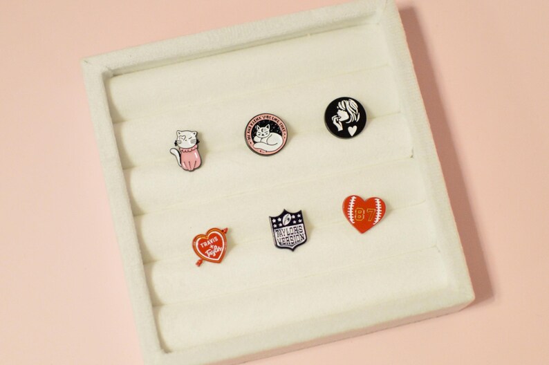 K&ouml;nnte beinhalten: Ein wei&szlig;es Tablett mit sechs Emaille-Pins. Die Pins zeigen eine Katze, eine schlafende Katze, ein Frauen Gesicht, ein Herz mit einem Pfeil, einen Footballhelm und ein Herz mit einem Football.
