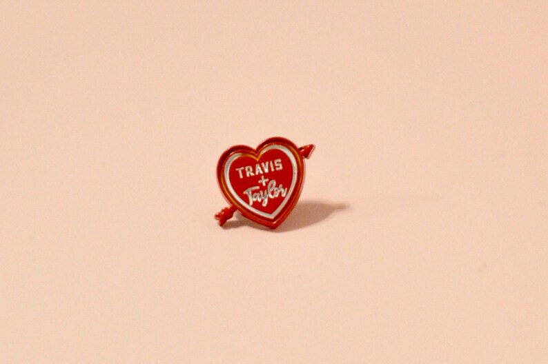 K&ouml;nnte beinhalten: Rote herzf&ouml;rmige Anstecknadel mit einem wei&szlig;en Pfeil und dem Text "TRAVIS + TAYLOR" in wei&szlig;en Buchstaben.