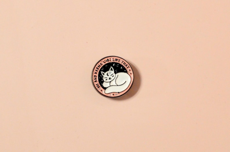 K&ouml;nnte beinhalten: Rosa emaillierte Anstecknadel mit einer schlafenden wei&szlig;en Katze in einem schwarzen Kreis. Der Text um den Kreis lautet "The and Karma Vibe Like That."