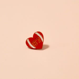 K&ouml;nnte beinhalten: Rote herzf&ouml;rmige Anstecknadel mit einem wei&szlig;en Baseball-Steppdesign und der Nummer "87" in goldener Schrift.