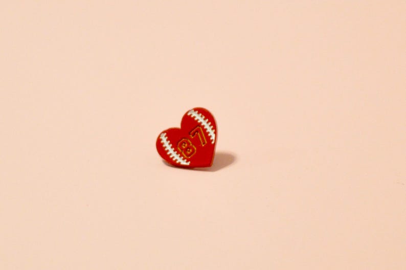 K&ouml;nnte beinhalten: Rote herzf&ouml;rmige Anstecknadel mit einem wei&szlig;en Baseball-Steppdesign und der Nummer "87" in goldener Schrift.