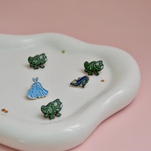 May include: Five enamel pins on a white ceramic plate. The pins are green frogs, a blue glass slipper, and a blue dress. The plate is shaped like a cloud.