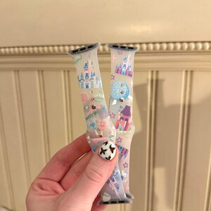 May include: Two clear Apple Watch bands with colorful Disney park designs. The bands feature images of Cinderella's Castle, Space Mountain, and other park icons.