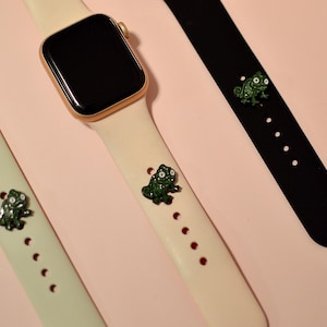 May include: Three Apple Watch bands with green chameleon charms. The bands are white, black, and light green.