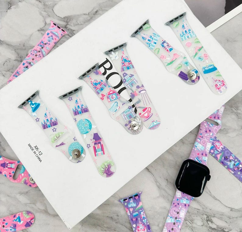 May include: A set of clear, colorful, silicone Apple Watch bands with a Disney castle and other theme park designs. The bands are on a white surface with the text "XK-12 MADE IN CHINA".