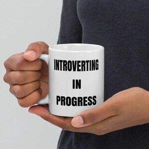 May include: White ceramic mug with the text "INTROVERTING IN PROGRESS" in bold black letters. The mug has a standard handle and is held by a person. The mug is a standard size for coffee or tea.