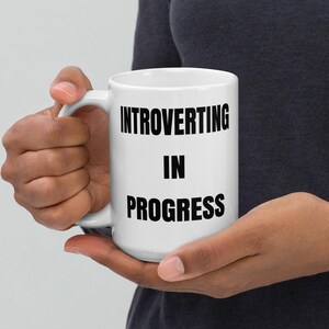 May include: White ceramic mug with the text "INTROVERTING IN PROGRESS" in black, held by a person. The mug has a standard handle and a glossy finish, suitable for hot or cold beverages. The mug is a standard 11 oz size.