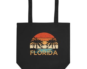 May include: Black tote bag with a retro sunset design featuring palm trees and the word "FLORIDA" in a vintage font. The design includes orange, yellow, and blue hues, creating a tropical feel. The bag has two black handles.