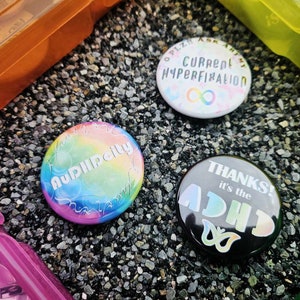 May include: Three round button pins with colorful designs. The first pin is rainbow colored with the text "AUDIIIPAILY" in white. The second pin is white with the text "PLZ ASK ABOUT MY CURRENT HYPERFIXATION" in black. The third pin is black with the text "THANKS! it's the ADHD" in rainbow colors.
