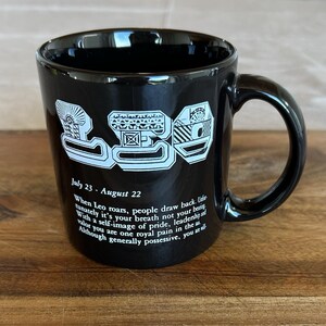 May include: A black ceramic mug featuring the word LEO in large white stylized letters at the top. Below the text, it reads July 23 - August 22, followed by a paragraph of text describing the Leo zodiac sign, including the phrase royal pain in the ass.
