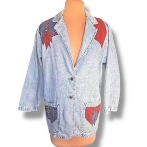 May include: Light blue denim jacket with decorative patches in red, blue, and black. The jacket has two front pockets and a button closure. The patches feature geometric and tribal-inspired designs. The jacket is displayed on a mannequin.