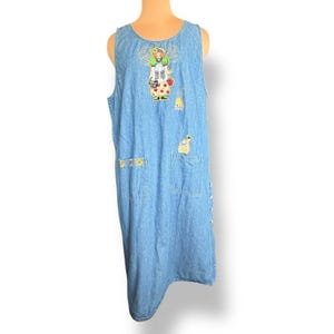 May include: Light blue denim dress with a sleeveless design. The dress features embroidered patches of a fairy, a bee, and other whimsical designs. It has two front pockets and decorative buttons on the side.
