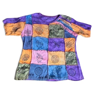 Könnte beinhalten: Ein Patchwork-T-Shirt in den Farbtönen Lila, Orange, Blau und Grün. Das Shirt hat eine Vielzahl von Blumen- und Spiraldesigns in jedem Quadrat. Das kurzärmelige Oberteil liegt flach auf einer weißen Oberfläche.