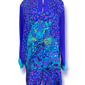 May include: A long-sleeved tunic in shades of purple and blue, featuring a large, stylized jaguar design on the front. The tunic has a button-up front and a round neckline. The sleeves are long with turquoise cuffs.