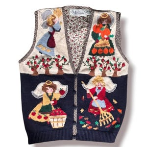 May include: A sleeveless knit vest with a V-neckline and button-front closure. The upper section is cream with two angels and apple trees, while the lower section is navy blue with two angels harvesting apples and raking leaves. Features a BellePointe label.