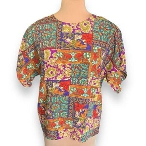 May include: A short-sleeved blouse featuring a vibrant patchwork pattern with rectangular blocks of floral and ornate motifs in shades of purple, teal, orange, red, and gold. The garment has a round neckline and a relaxed, boxy fit.