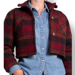 Puede incluir: Chaqueta corta a cuadros rojos, negros y marrones con cuello y dos botones grandes. La chaqueta se lleva sobre una camisa vaquera azul claro. La chaqueta tiene dos bolsillos delanteros.