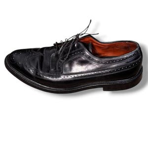 May include: Black leather dress shoe with brogue detailing and a lace-up closure. The shoe features a classic wingtip design with decorative perforations along the edges and a polished finish. The interior lining is a warm brown color.