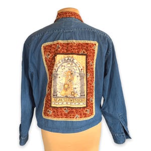 May include: Blue denim jacket with a decorative back panel. The panel features a detailed illustration with the words "GRACE" and "BEAUTY WISDOM" in a rectangular frame with a red and gold pattern. The jacket has long sleeves and a button-down front.