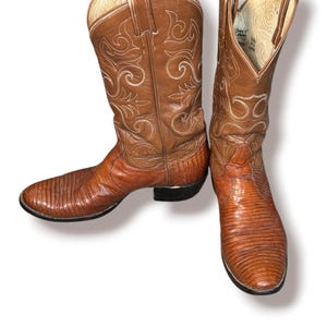 May include: A pair of brown leather cowboy boots featuring a textured lizard-skin pattern on the foot and a smooth leather shaft with intricate white stitching. The boots have pull straps at the top and a stacked heel with a black sole.