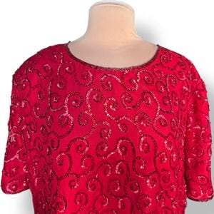 May include: A vibrant red top with short sleeves, adorned with intricate sequin and bead embellishments in a swirling pattern. The neckline is also embellished with beads. The top is displayed on a mannequin.