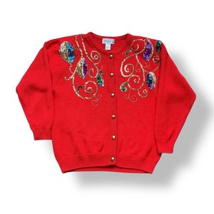 May include: A red cardigan sweater with gold buttons and embellished with colorful jewels and gold sequins. The sweater has a round neckline and long sleeves. The embellishments are arranged in a swirling pattern across the shoulders and chest.