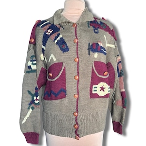 Könnte beinhalten: Ein beigefarbener Cardigan mit Knopfleiste, verziert mit farbenfrohen Strickmustern zum Thema Flugzeuge und Militär. Der Pullover hat zwei Vordertaschen mit bordeauxfarbenem Besatz und Holzknöpfen. Die Manschetten und der untere Saum sind bordeauxfarben.