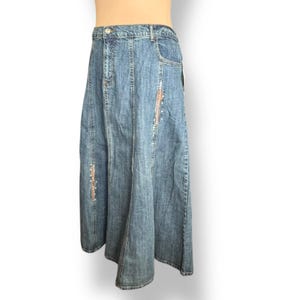 May include: A long blue denim skirt featuring a high-waisted design with a button and zipper closure. The skirt has vertical panel seams and two vertical decorative strips with small metallic studs along the front side panels.