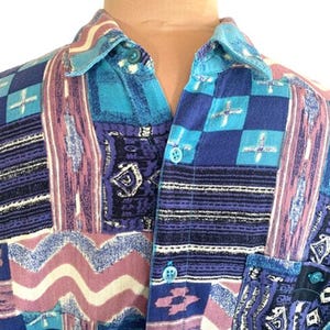May include: A button-down shirt featuring a vibrant patchwork pattern in shades of blue, purple, and white. The design includes geometric shapes, wavy lines, and intricate motifs. The shirt has a classic collar and matching blue buttons down the front.