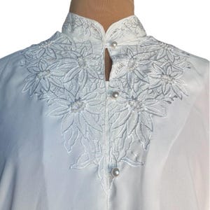 May include: White blouse with a mandarin collar and floral embroidery. The embroidery features a detailed design of flowers and leaves, accented with small pearls. The blouse has a button closure and a high neckline.