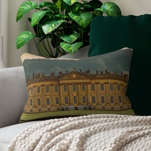 Puede incluir: Un cojín rectangular decorativo con una ilustración vintage de un gran edificio con columnas y ventanas. El cojín está colocado sobre un sofá gris con otros cojines y una manta crema. Una planta verde está en el fondo.