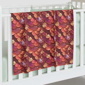 May include: A red, gold, and pink glittery mermaid scale patterned baby blanket. The blanket is folded and draped over a white crib.