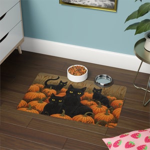 Peut inclure: Un tapis pour animaux de compagnie sur le thème du chat noir avec un fond brun et des citrouilles orange. Quatre chats noirs sont assis sur les citrouilles. Le tapis est conçu pour les bols de nourriture et d'eau pour animaux de compagnie.