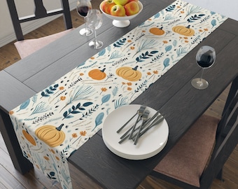 May include: A white table runner with a pattern of orange pumpkins, blue leaves, and brown stems on a cream background. The runner is laid on a dark wood table with a plate and cutlery.