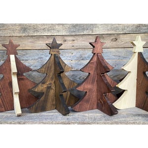 May include: Four wooden Christmas trees in varying shades of brown and natural wood. Each tree has a star on top. The trees are displayed against a rustic wooden background, showcasing a festive, handcrafted aesthetic.