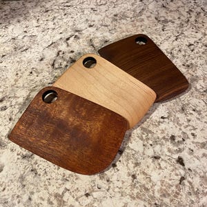 Peut inclure: Trois planches à découper en bois de différentes nuances de brun. Chaque planche a une forme arrondie avec un trou pour suspendre. Le grain du bois est visible, mettant en valeur la beauté naturelle du matériau.