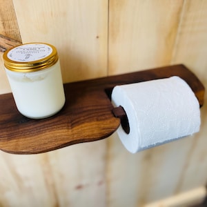 Peut inclure: Un porte-papier toilette en bois brun foncé avec une étagère à bord naturel. Une bougie blanche dans un pot en verre avec un couvercle doré est posée sur le côté gauche de l'étagère, tandis qu'un rouleau de papier toilette blanc est fixé à droite.