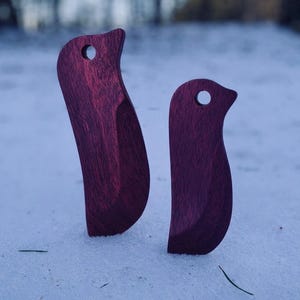 May include: Two dark purple wooden bird-shaped ornaments with smooth, curved silhouettes and a single circular hole near the top of each piece. The wood grain is visible throughout the deep violet surface of both carved figures.