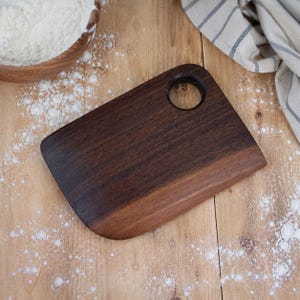 May include: A rectangular dark brown wooden chopping board with rounded corners and a circular hole near the top right corner. The board features a smooth, polished surface with visible wood grain patterns and a slightly lighter tone at the bottom edge.