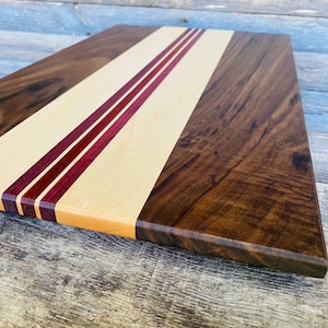 May include: A rectangular wooden cutting board with a light wood center stripe and two parallel burgundy stripes. The sides are dark brown wood. The board is approximately 16 inches long and 10 inches wide.