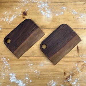 May include: Two rectangular dark brown wooden cutting boards with rounded corners and a circular hole on one side. The boards feature a visible wood grain pattern and are positioned at an angle on a light-colored wooden surface with white powder.