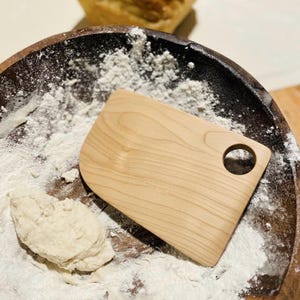 May include: A light-coloured wooden dough scraper with a round handle hole rests in a dark bowl dusted with flour. A small portion of dough sits near the scraper, suggesting baking or cooking.